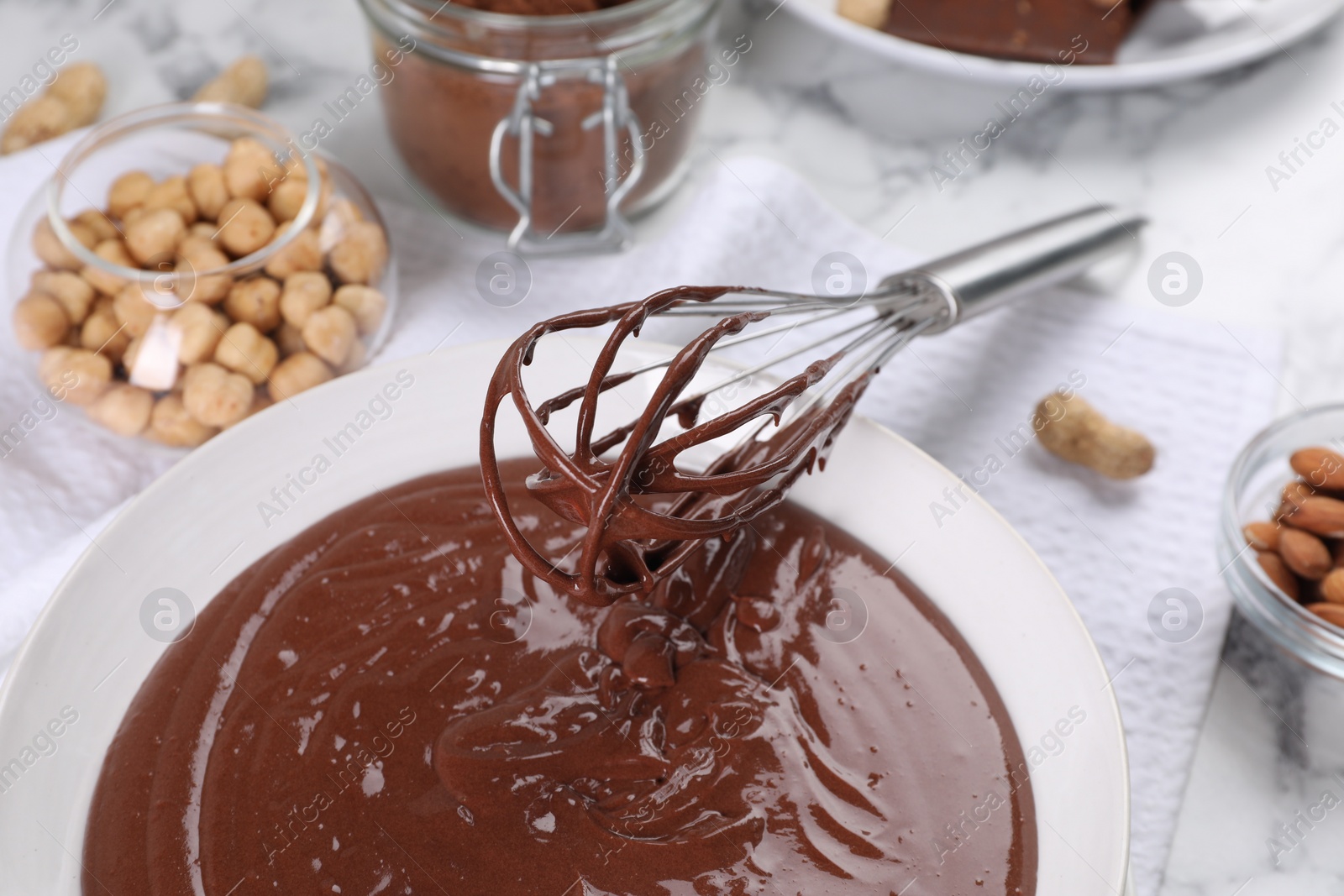 Photo of Bowl of chocolate cream, whisk, and nuts on white marble table, closeup