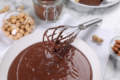 Photo of Bowl of chocolate cream, whisk, and nuts on white marble table, closeup