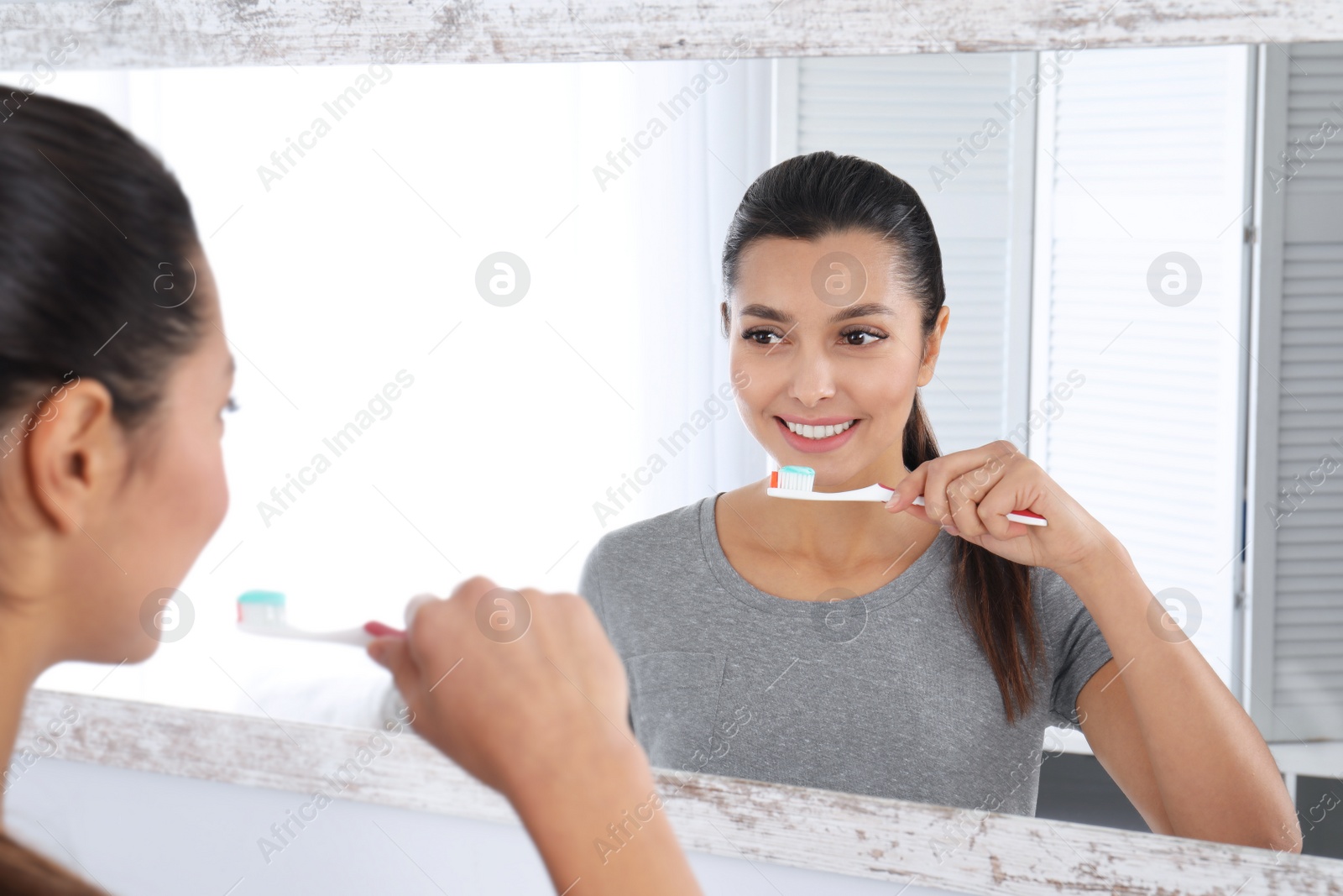 Photo of Young woman cleaning teeth against mirror in bathroom