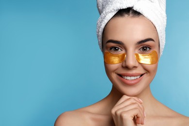 Beautiful young woman with under eye patches and hair wrapped in towel on light blue background, space for text