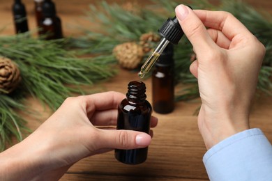Photo of Woman holding pipette with pine essential oil over bottle at wooden table, closeup