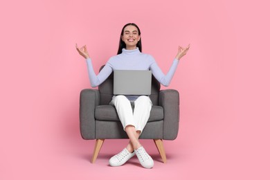 Happy woman with laptop sitting in armchair and meditating on pink background