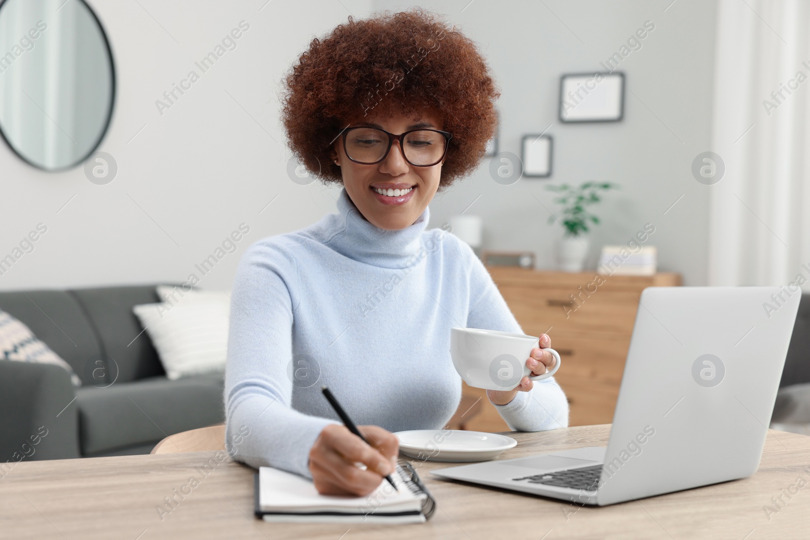 Photo of Beautiful young woman writing in notebook near laptop at wooden desk in room