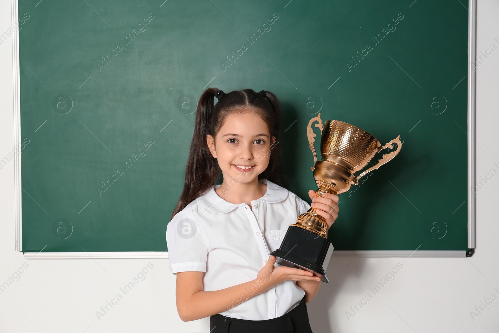 Photo of Happy girl with golden winning cup near chalkboard in classroom
