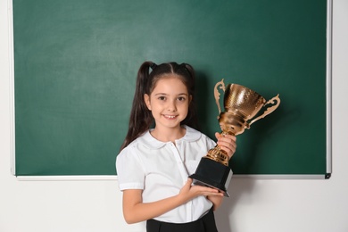 Happy girl with golden winning cup near chalkboard in classroom