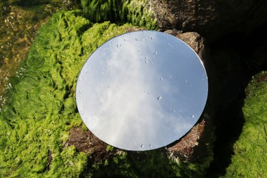 Round mirror reflecting light blue sky on stones with seaweed outdoors