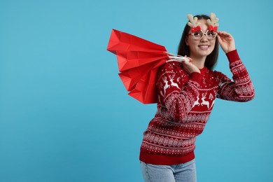 Photo of Happy young woman in Christmas sweater and funny glasses with shopping bags on light blue background. Space for text
