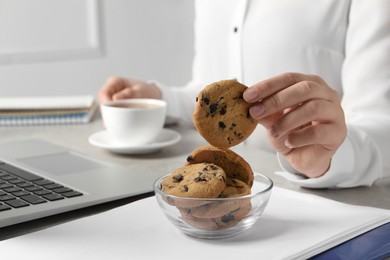 Photo of Woman taking chocolate chip cookie from bowl while drinking tea in office, closeup