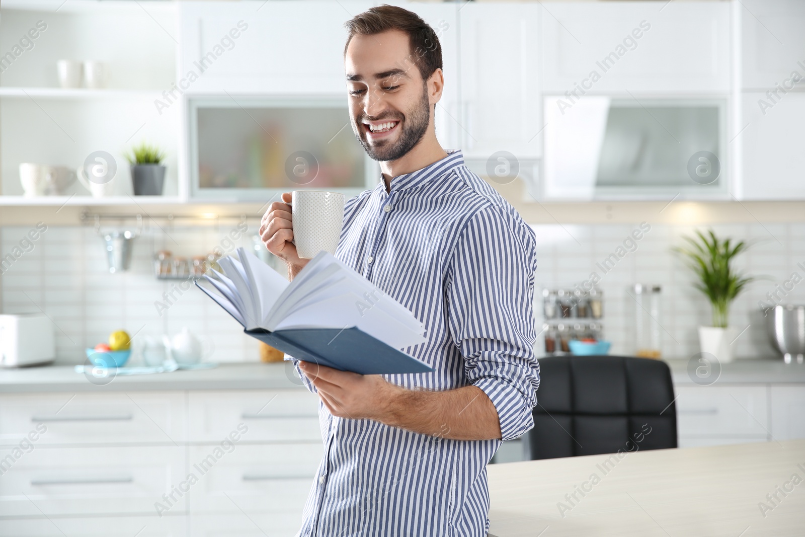 Photo of Handsome young man with cup of coffee reading book at home