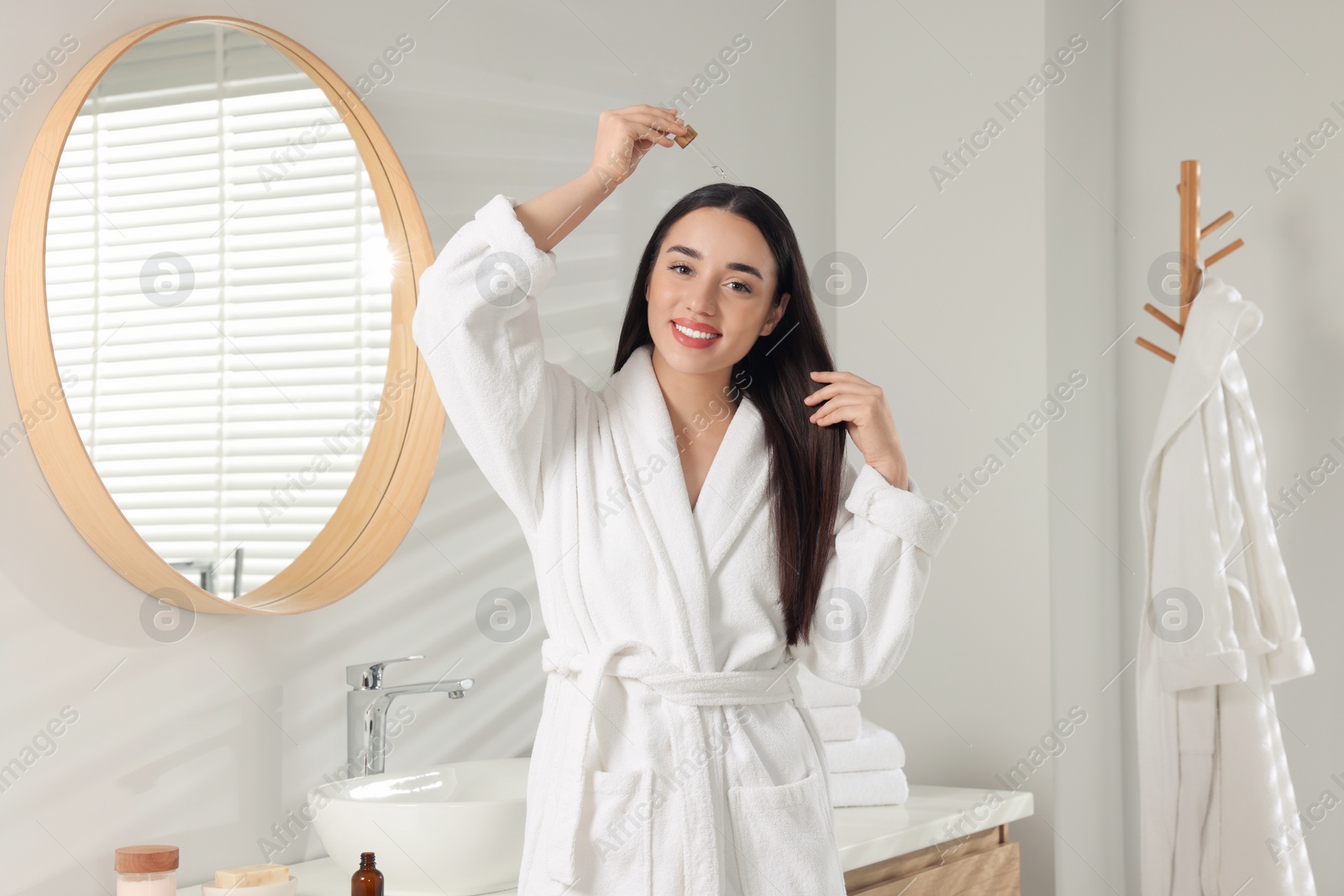 Photo of Happy young woman applying essential oil onto hair roots in bathroom