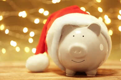 Photo of Piggy bank with Santa hat on table against blurred Christmas lights. Space for text