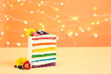 Photo of Slice of delicious rainbow cake with flowers on table against blurred lights