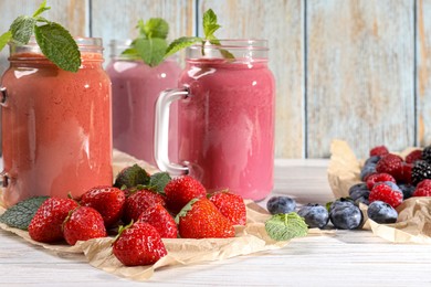 Photo of Mason jars of different berry smoothies and fresh ingredients on white wooden table