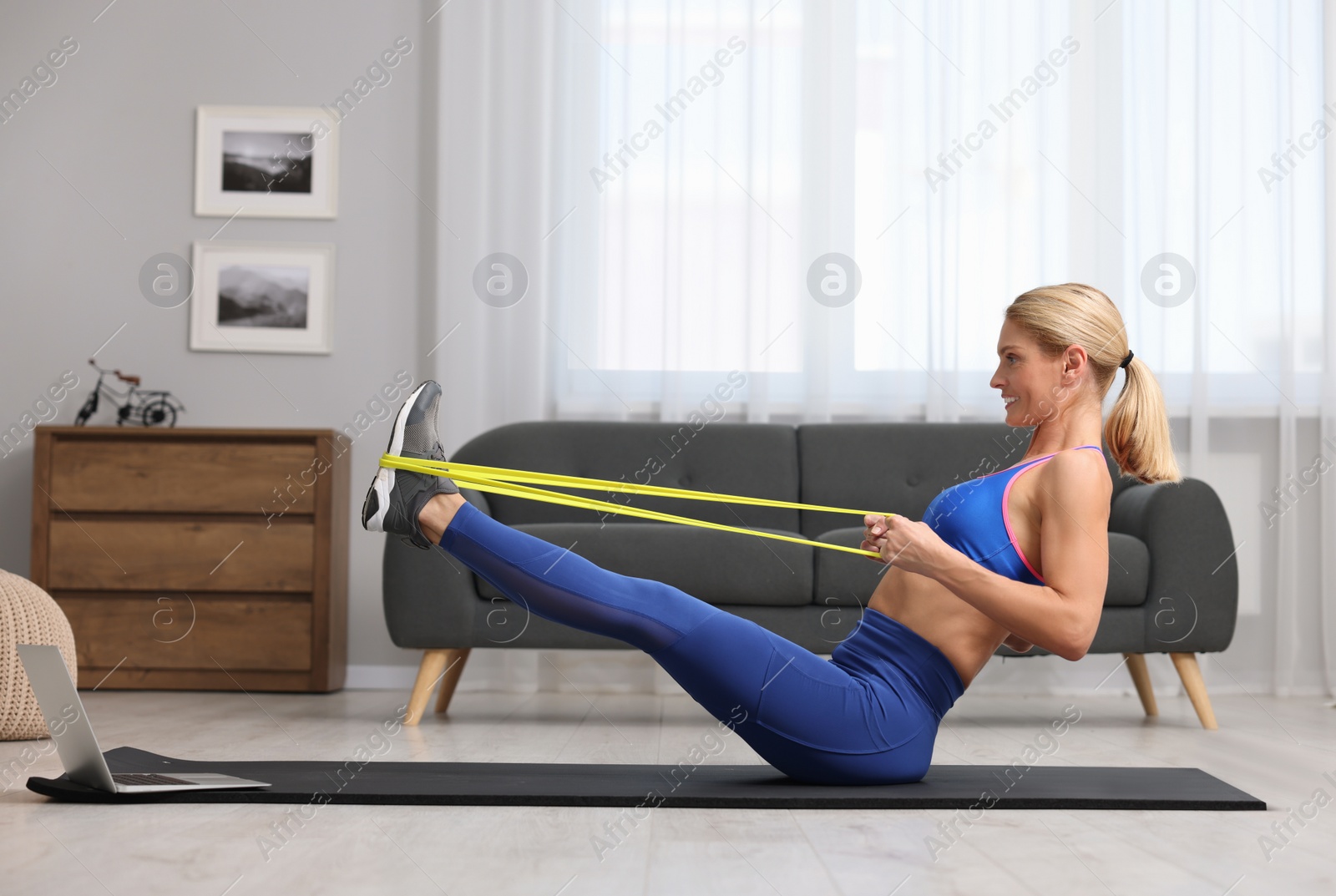 Photo of Fit woman doing exercise with fitness elastic band near laptop on mat at home