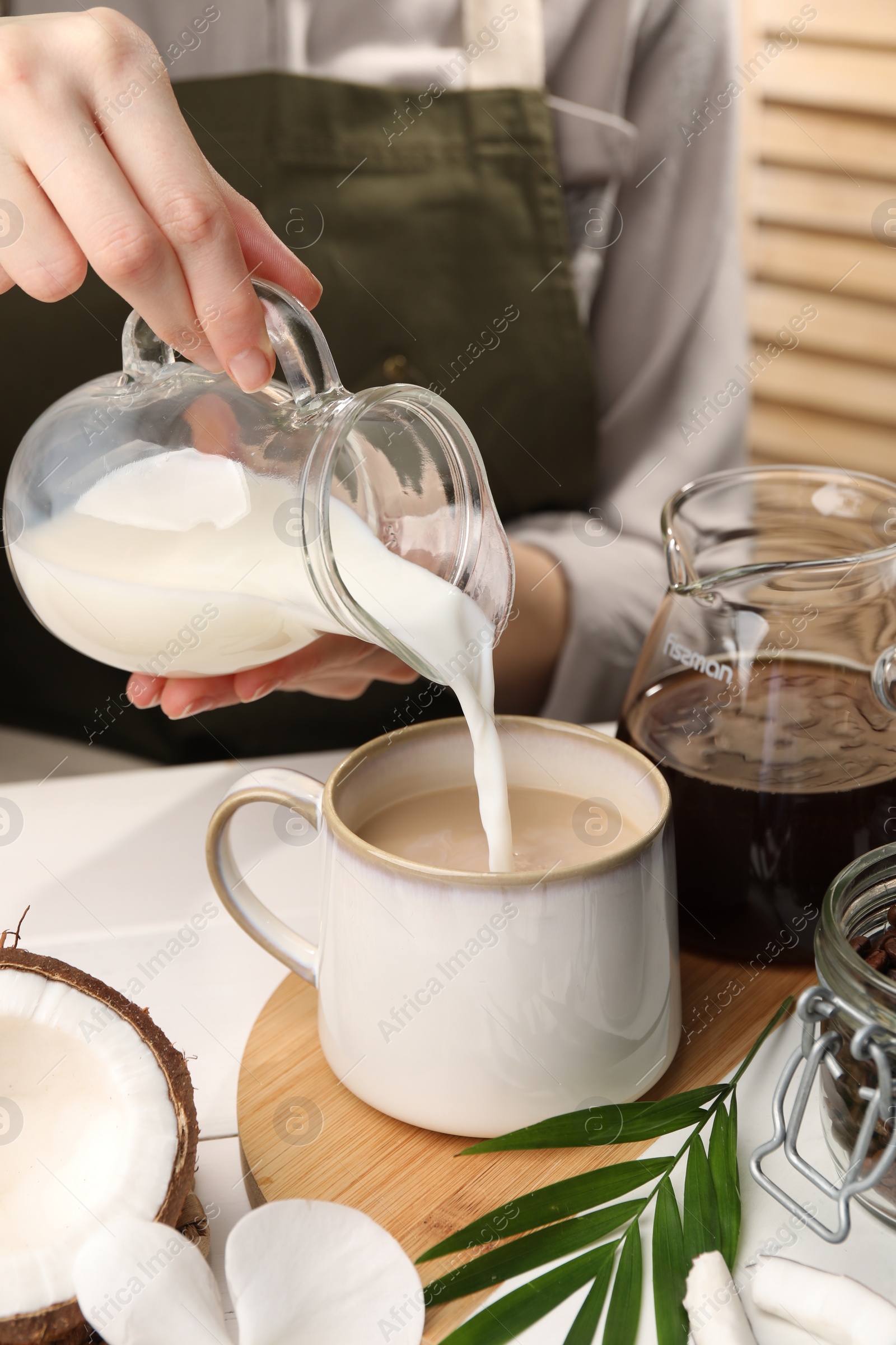 Photo of Woman pouring tasty coconut milk into mug of coffee at white table indoors, closeup