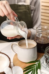 Woman pouring tasty coconut milk into mug of coffee at white table indoors, closeup