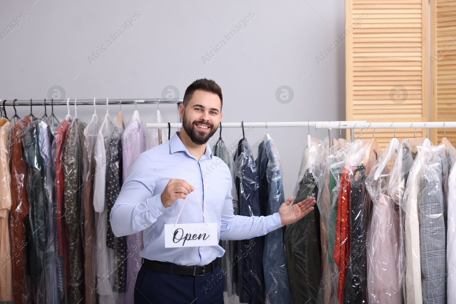 Photo of Dry-cleaning service. Happy worker holding Open sign near racks with clothes indoors