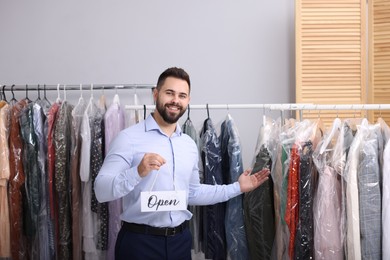 Dry-cleaning service. Happy worker holding Open sign near racks with clothes indoors