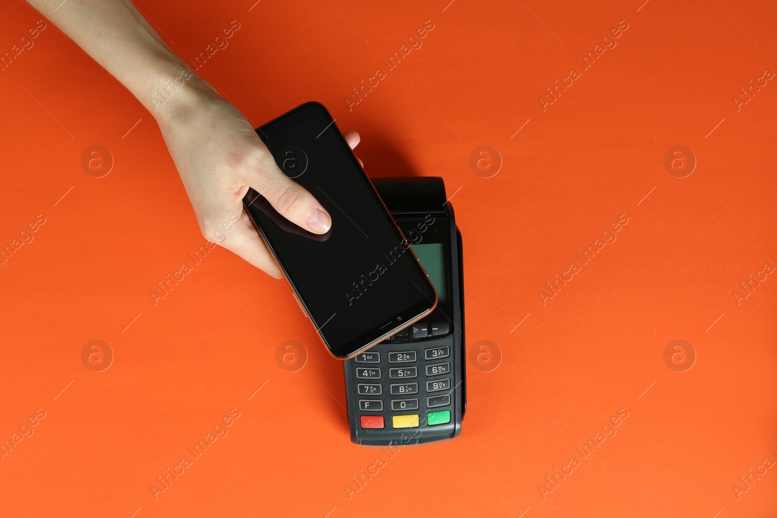 Photo of Woman with smartphone using modern payment terminal on orange background, top view