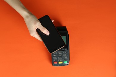 Photo of Woman with smartphone using modern payment terminal on orange background, top view