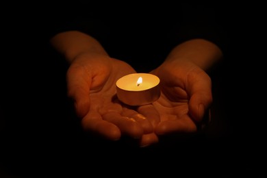 Woman holding burning candle in hands on black background, closeup