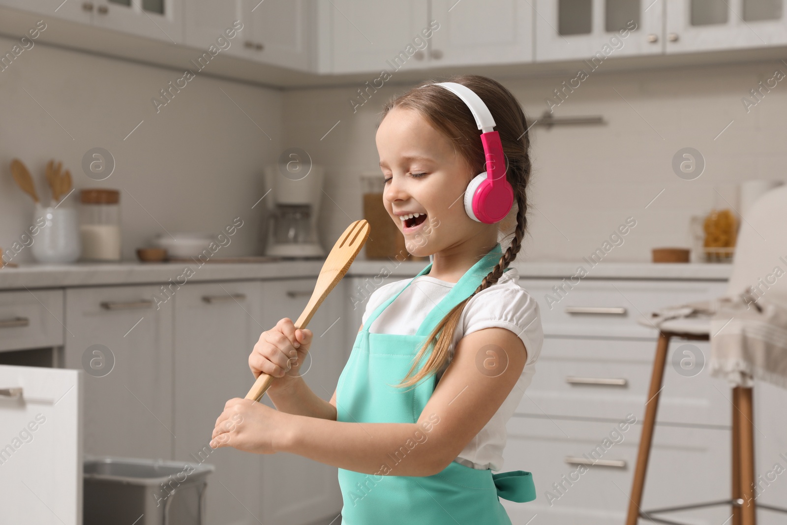 Photo of Cute little girl with headphones and fork spatula singing in kitchen