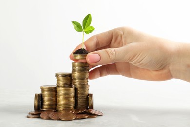 Woman putting coin onto stack with green plant on white marble table, closeup. Investment concept