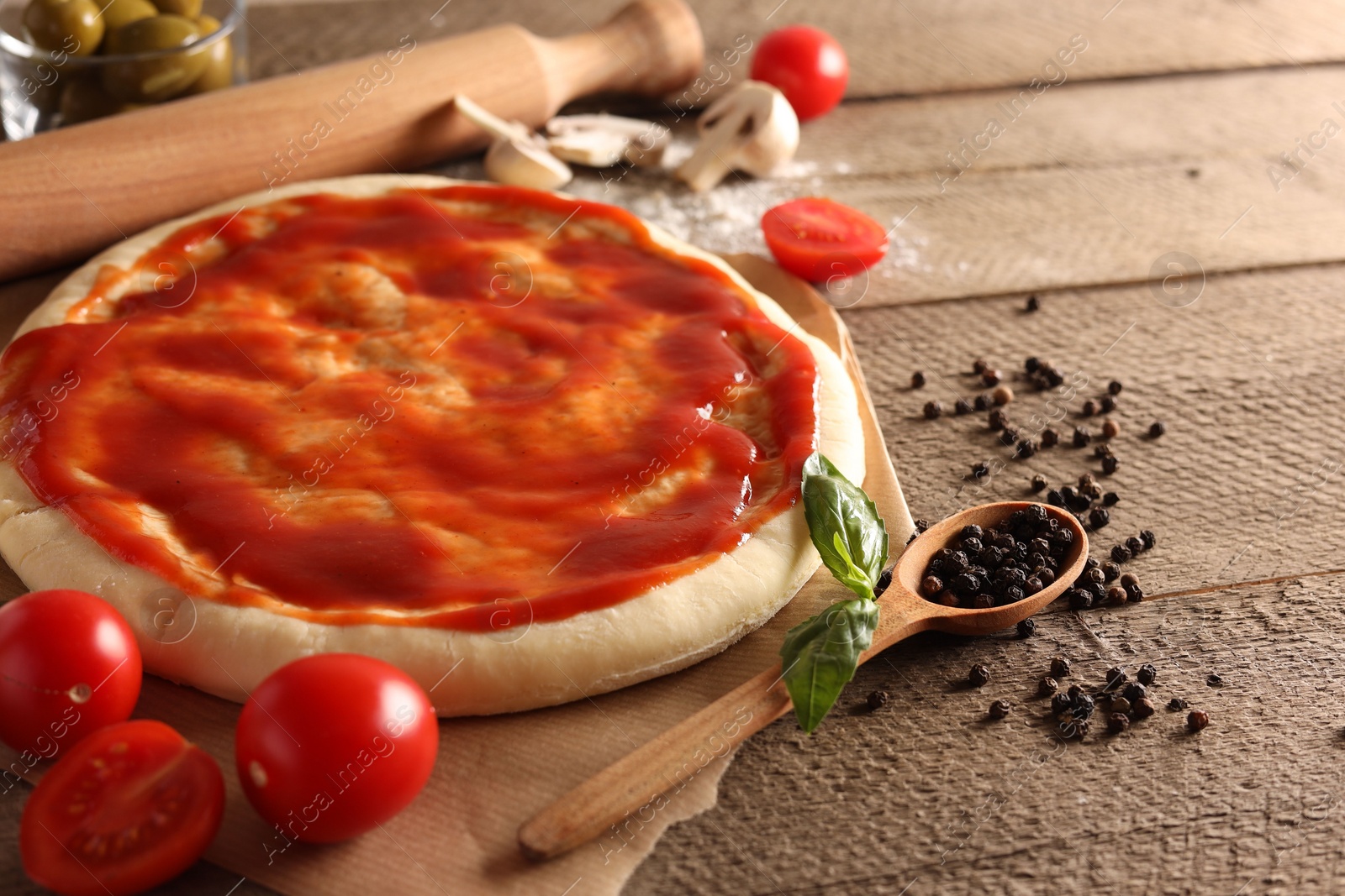 Photo of Pizza dough smeared with tomato sauce and products on wooden table, closeup