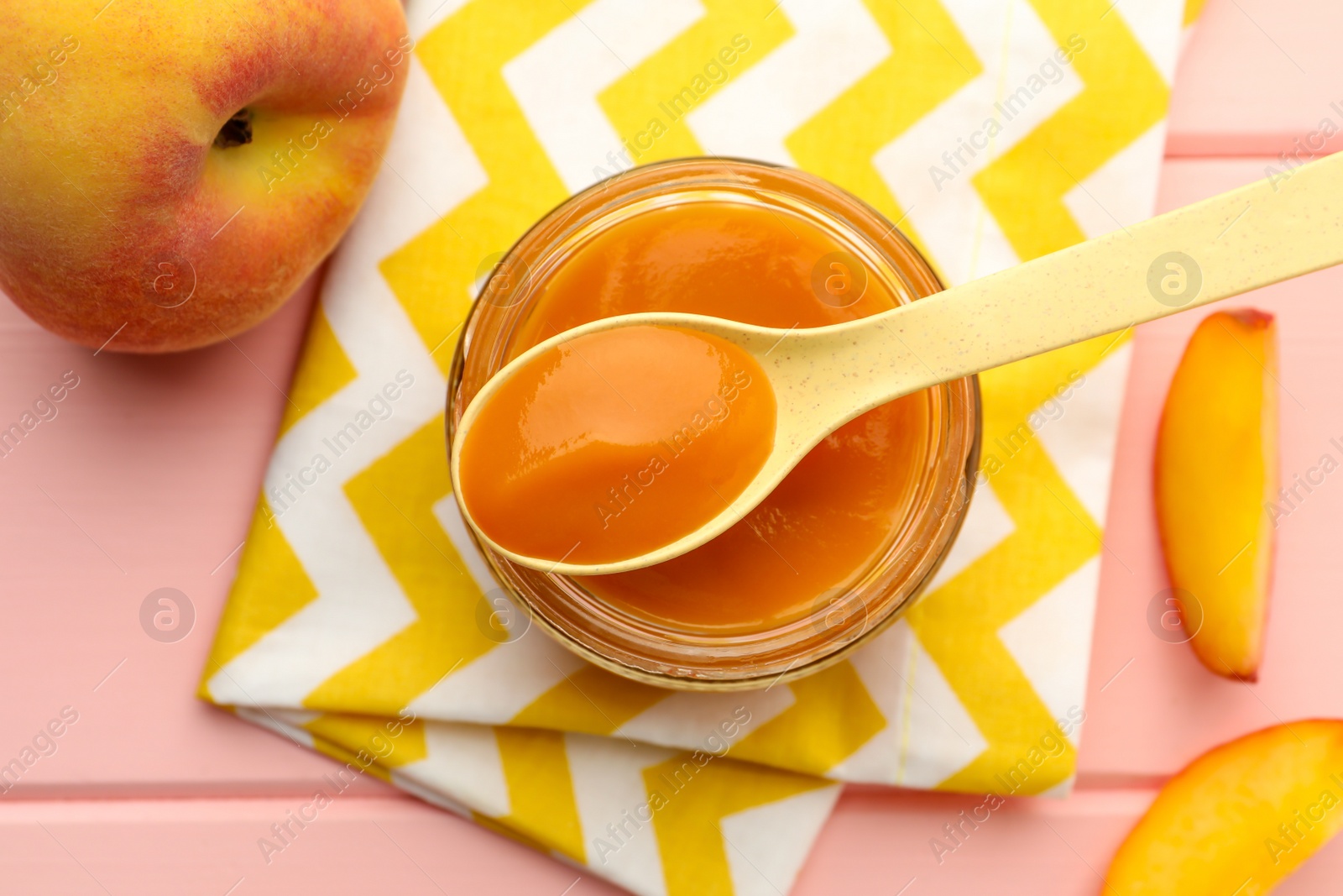 Photo of Spoon with healthy baby food over glass jar and fresh peaches on pink wooden table, flat lay