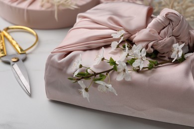 Photo of Furoshiki technique. Gift packed in pink fabric, beautiful flowers and scissors on white table, closeup
