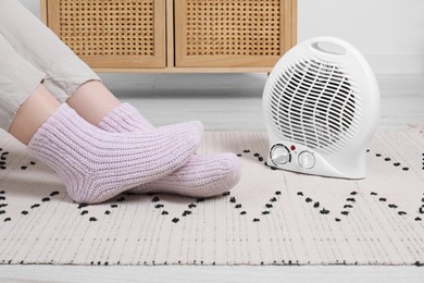 Photo of Woman warming feet near electric fan heater at home, closeup