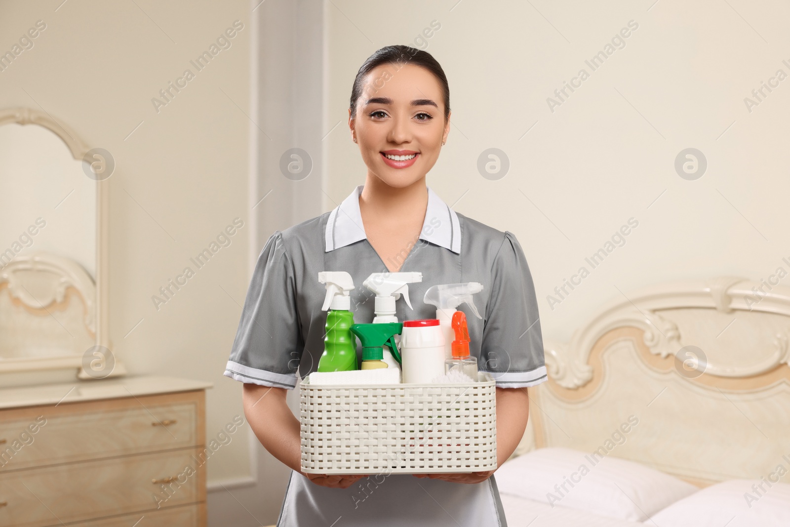 Photo of Young chambermaid holding basket with cleaning products in hotel room