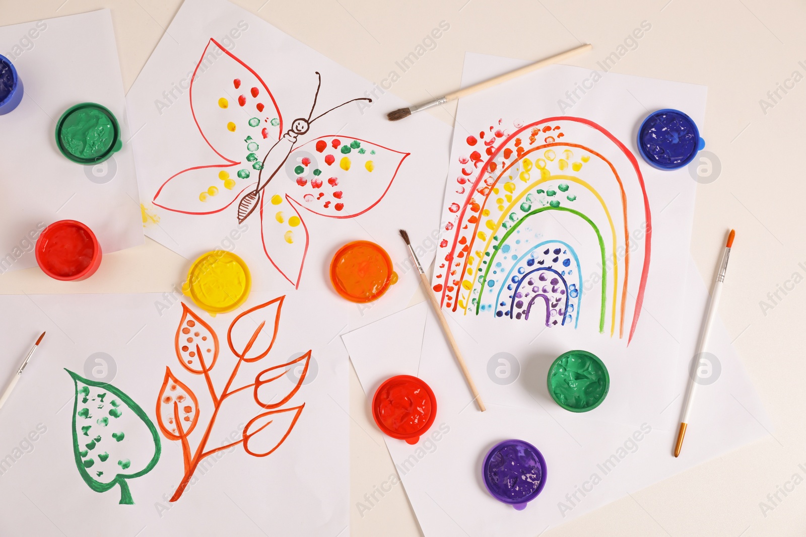 Photo of Cute children's drawings, brushes and set of paints on white table, flat lay