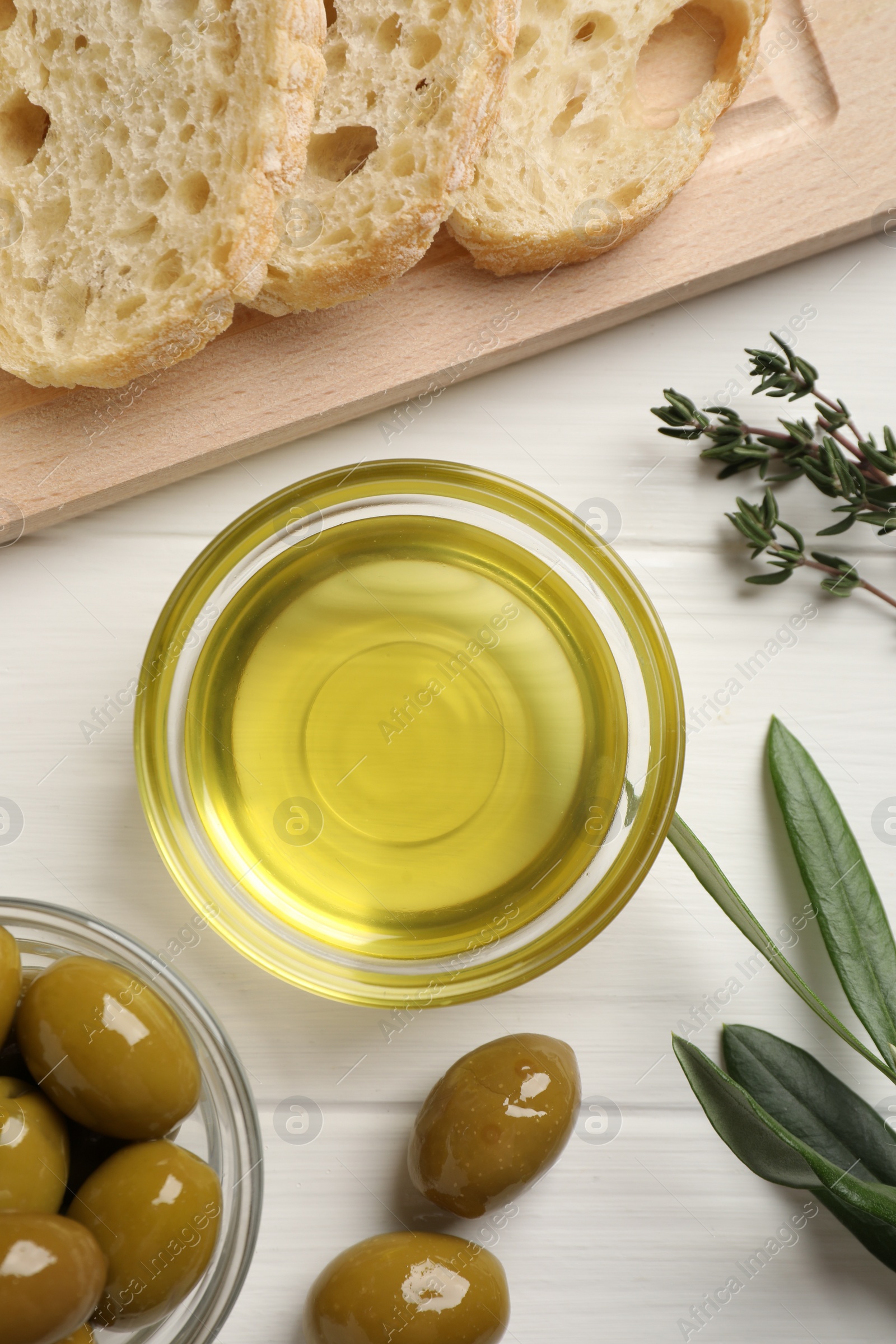 Photo of Bowl of cooking oil, ripe olives and bread on white wooden table, flat lay