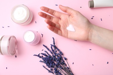 Woman applying hand cream and lavender flowers on pink background, top view