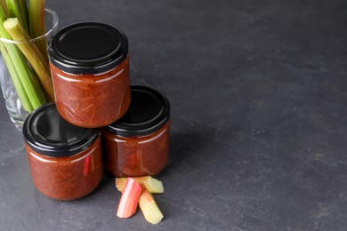Photo of Jars of tasty rhubarb jam and stalks on grey table. Space for text