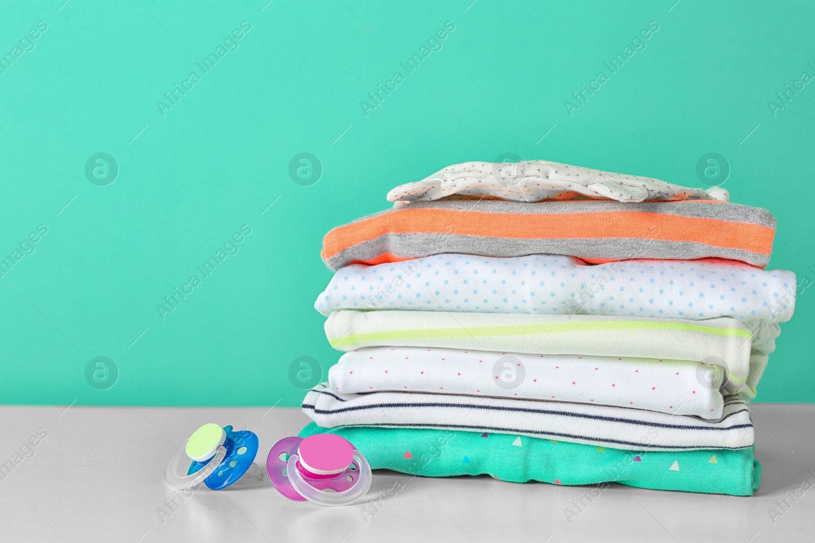 Photo of Stack of stylish child clothes and pacifiers on table against color background