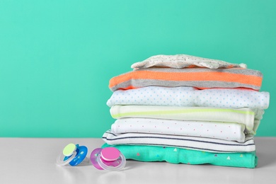 Stack of stylish child clothes and pacifiers on table against color background