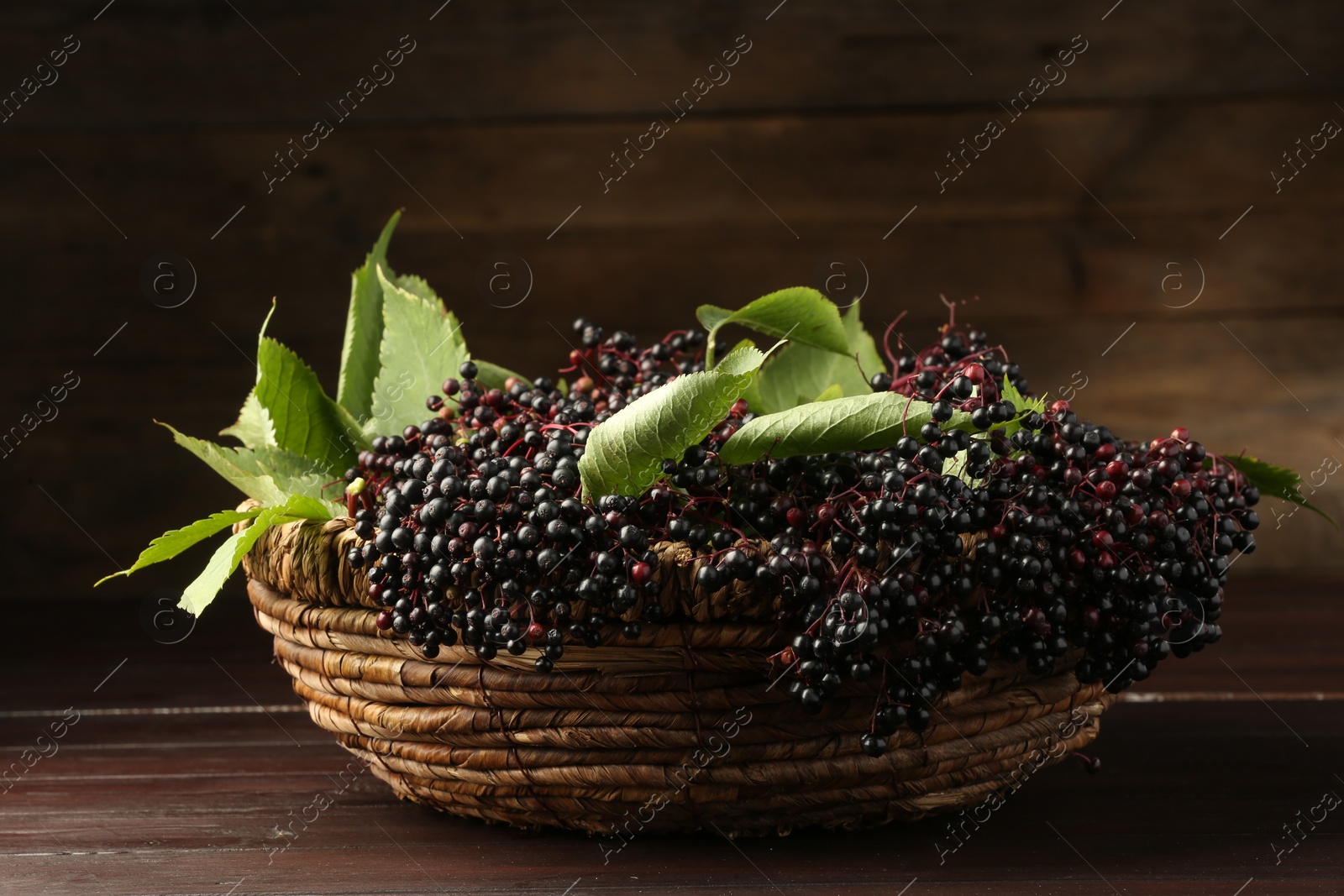 Photo of Ripe elderberries with green leaves in wicker basket on wooden table