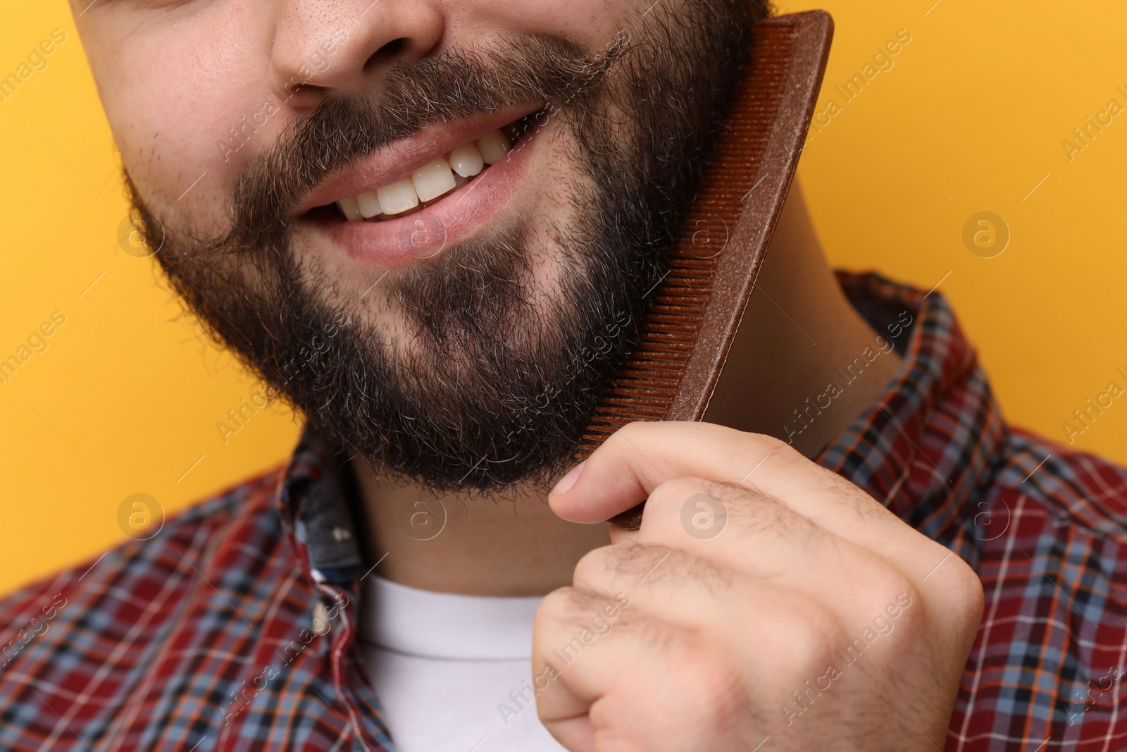 Photo of Handsome young man combing beard on yellow background, closeup