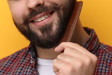 Handsome young man combing beard on yellow background, closeup