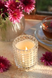 Photo of Burning scented candle in glass holder and chrysanthemum flowers on beige textured table. Autumn season