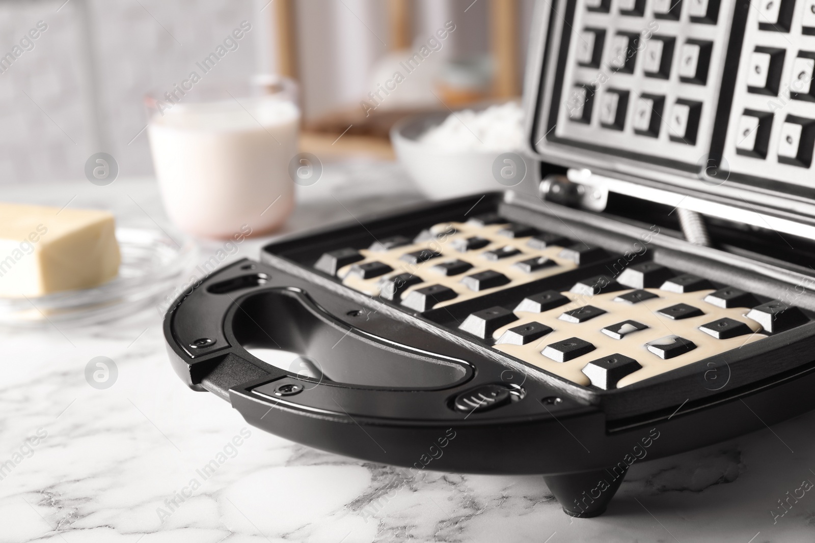 Photo of Belgian waffle maker with dough on white marble table, closeup