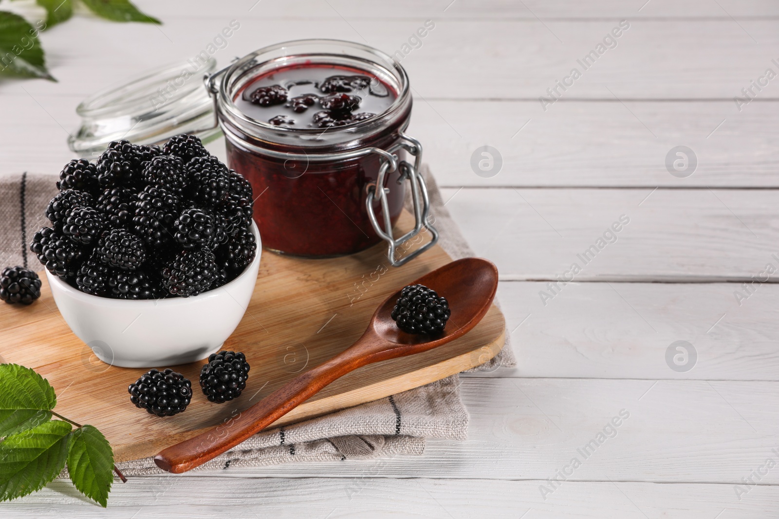 Photo of Fresh ripe blackberries, tasty jam and leaves on white wooden table. Space for text