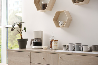 Photo of Modern coffeemaker and cups on commode near light wall indoors
