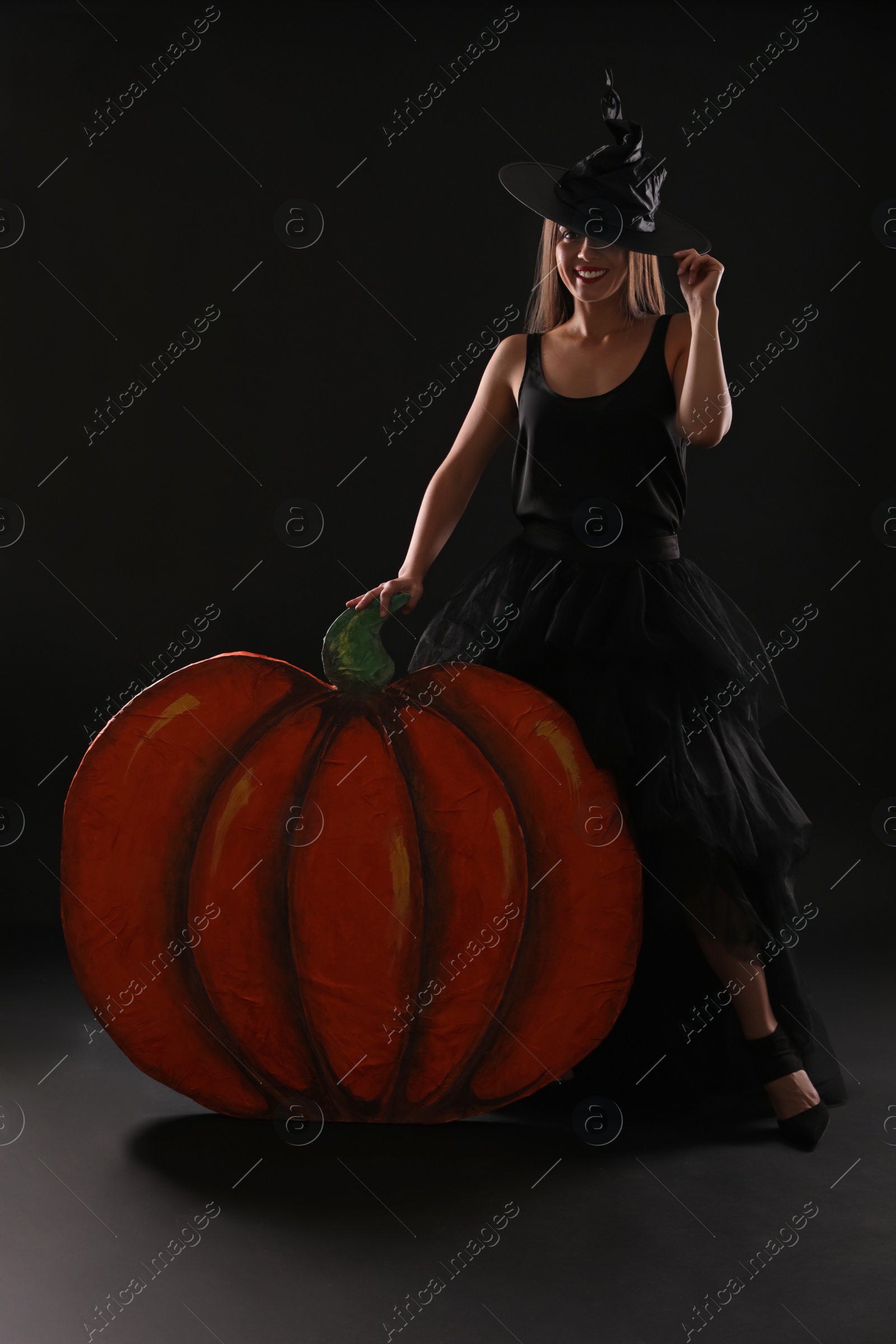 Photo of Young woman wearing witch costume near decorative pumpkin on black background. Halloween party