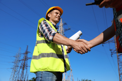 Photo of Professional electricians shaking hands near high voltage tower, closeup
