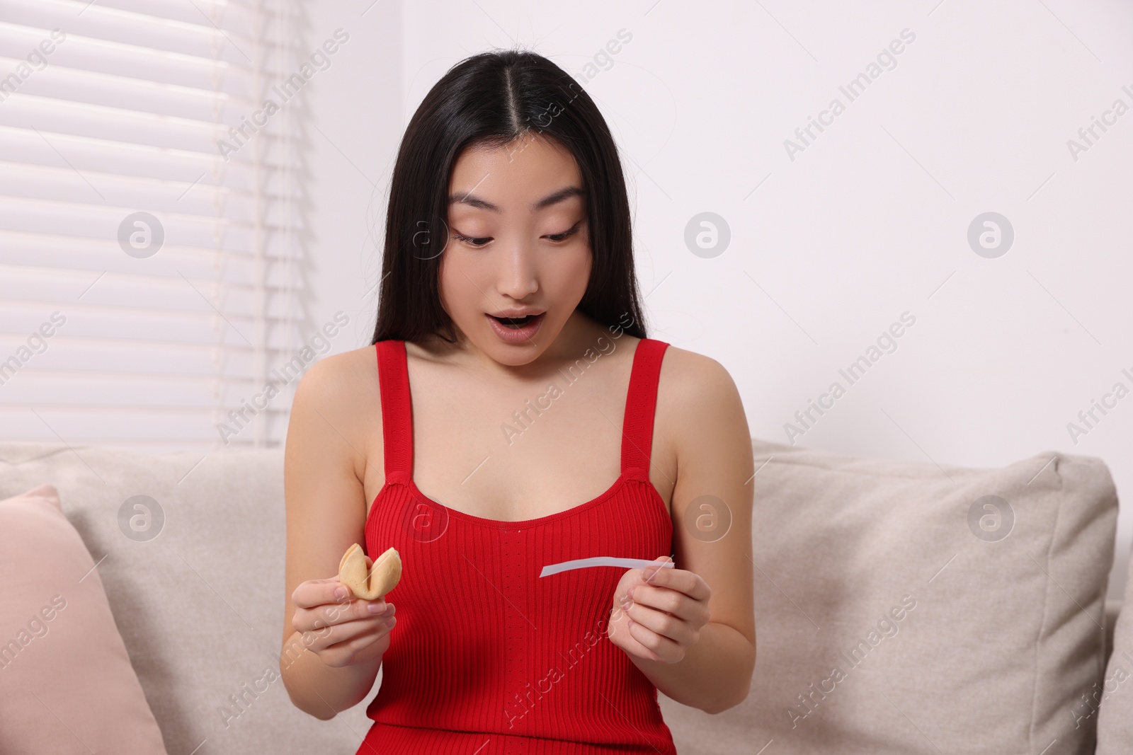 Photo of Surprised asian woman holding tasty fortune cookie with prediction on sofa at home