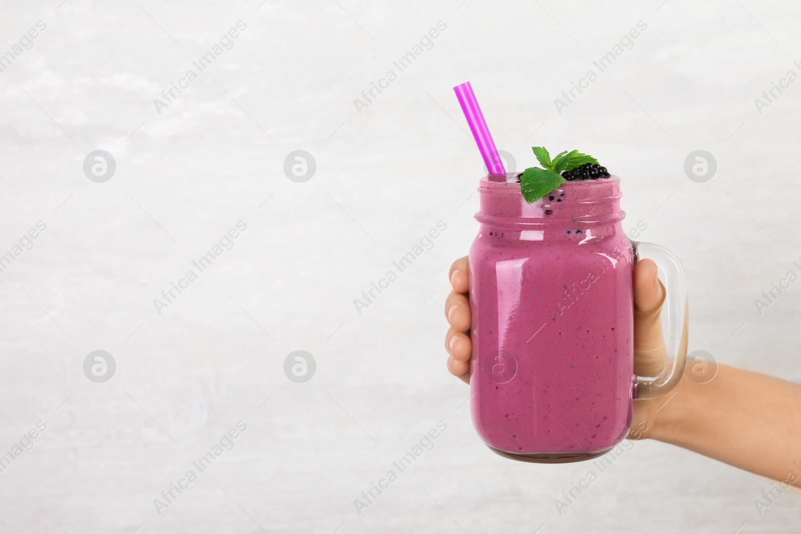 Photo of Young woman holding mason jar of tasty blackberry smoothie on light grey background, closeup. Space for text