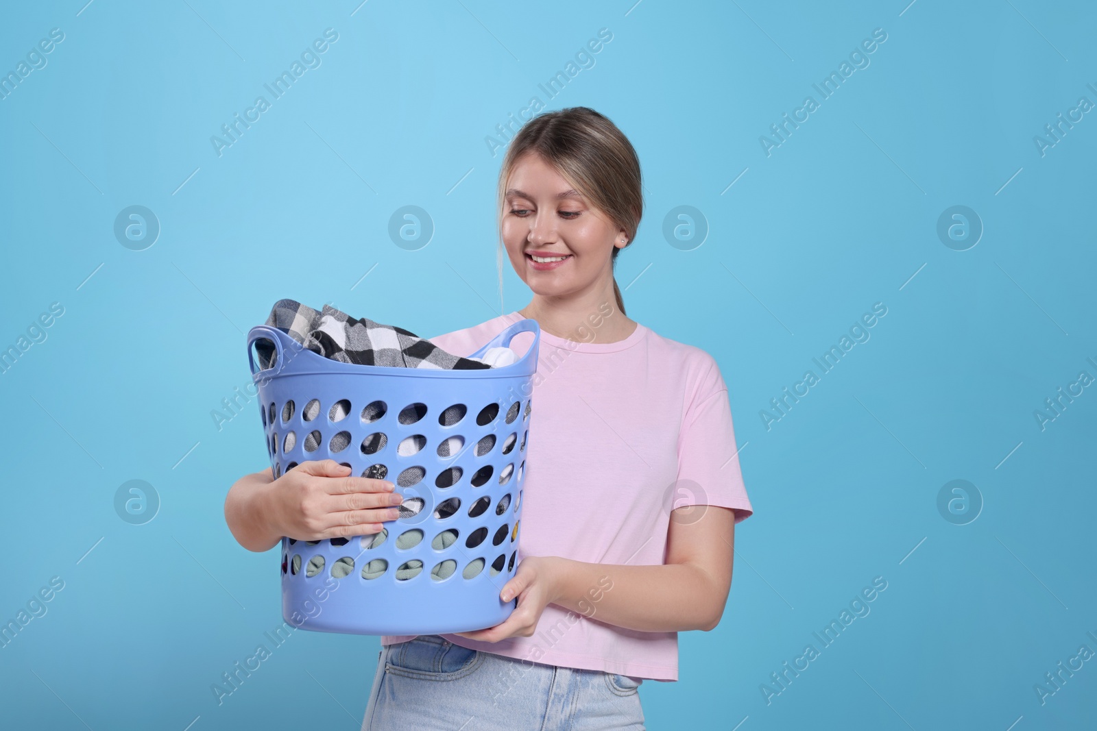 Photo of Happy woman with basket full of laundry on light blue background